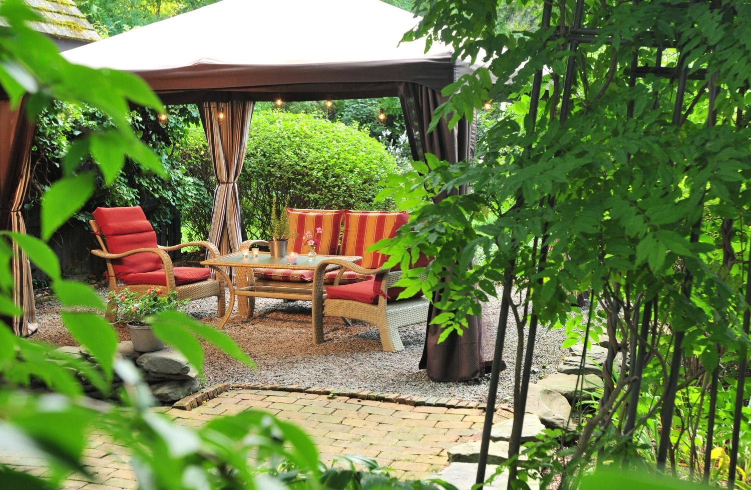 A gazebo with a couch and chairs underneath it in a garden surrounded by trees.